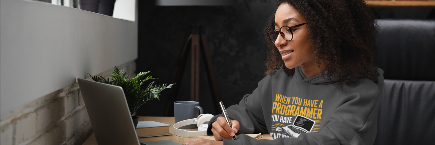 Person sitting at a desk with a laptop, pen, and coffee cup, wearing a gray sweatshirt with text.